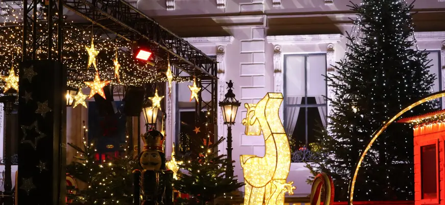 Escena nocturna navideña con decoración iluminada en forma de caballo de ajedrez, árboles de Navidad, estrellas doradas, cascanueces y farolas frente a la fachada de un edificio elegante.