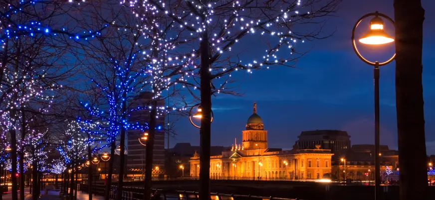 Vista nocturna del edificio histórico Custom House iluminado en Dublín, con árboles decorados con luces azules y blancas junto al río y farolas encendidas en el paseo.
