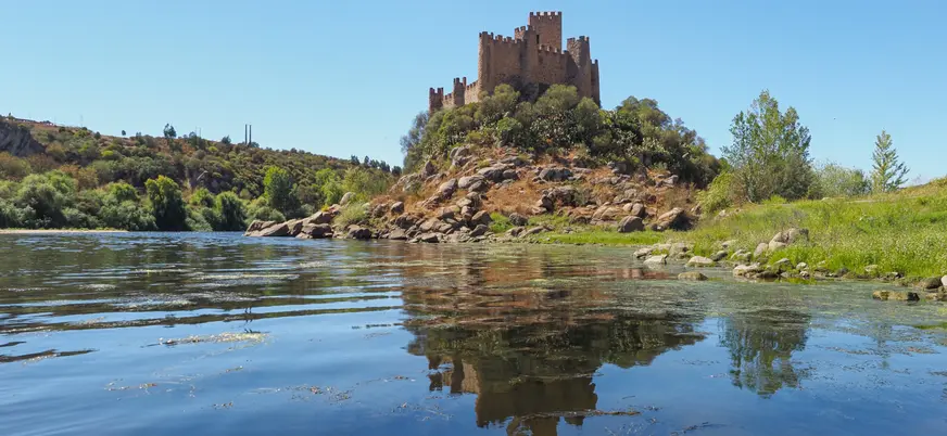 Castillo de Almourol reflejado en el río desde la orilla, bajo un cielo despejado.