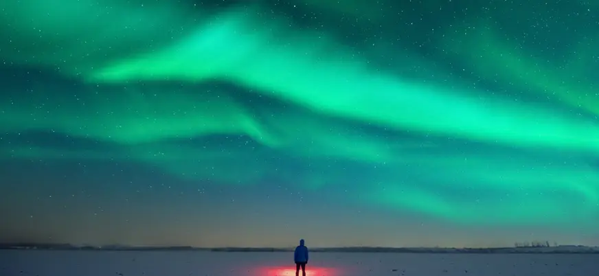 Persona de pie sobre nieve iluminada por luz roja, observando auroras boreales verdes que cubren el cielo estrellado.