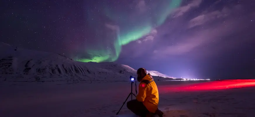 Persona con chaqueta amarilla fotografiando auroras boreales verdes sobre montañas nevadas, bajo un cielo estrellado con tonos púrpura.