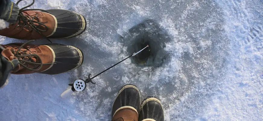 Dos pares de botas sobre hielo junto a un agujero para pesca en un lago congelado, con caña de pescar apoyada en el borde, en un paisaje invernal.