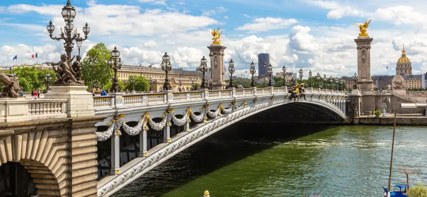 Puente Alexandre III sobre el río Sena en París, adornado con faroles y esculturas doradas, con cielo azul y nubes al fondo.