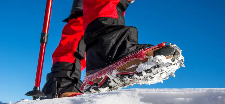 Primer plano de una bota con crampones sobre nieve, junto a un bastón de senderismo, con cielo azul despejado al fondo.