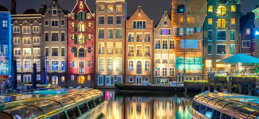 Fachada de casas estrechas y coloridas iluminadas por la noche, reflejadas en el canal de Ámsterdam, con barcos turísticos atracados en primer plano.