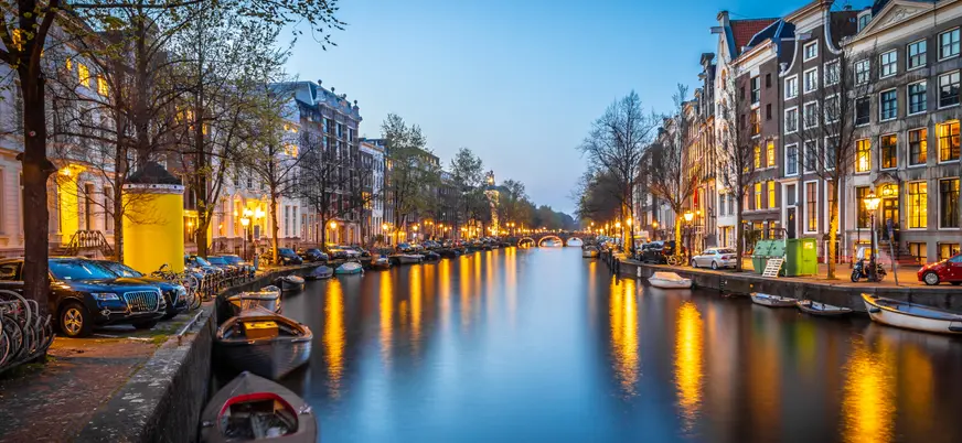 Canal de Ámsterdam al atardecer, bordeado por casas típicas neerlandesas, árboles y pequeñas embarcaciones, con luces reflejadas en el agua.
