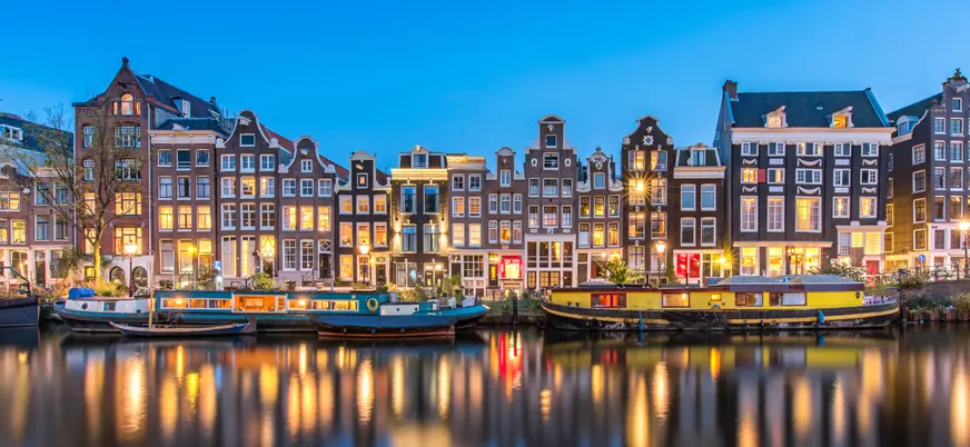 Casas tradicionales neerlandesas iluminadas al anochecer junto a un canal de Ámsterdam, con barcos estacionados y reflejos de los edificios en el agua.