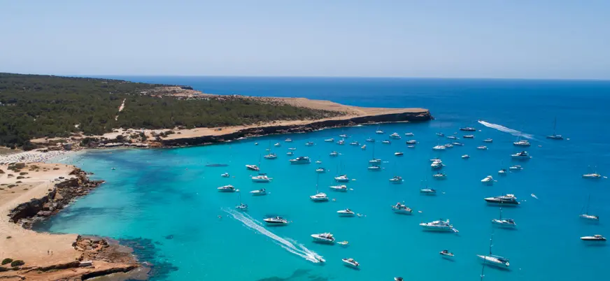 Cala Saona vista desde lo alto, con barcos, vegetación y aguas de un azul intenso.