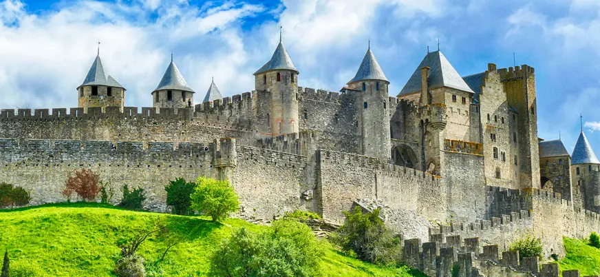 Vista de las murallas y torres medievales de Carcasona entre colinas verdes, bajo un cielo dramático.