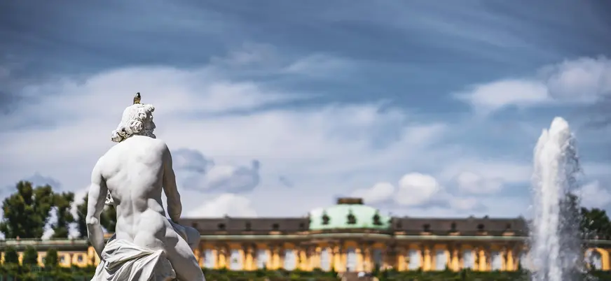 Estatua clásica con un pájaro en el Jardín de Sanssouci, Potsdam. Al fondo se ve la fuente y el Palacio de Sanssouci.