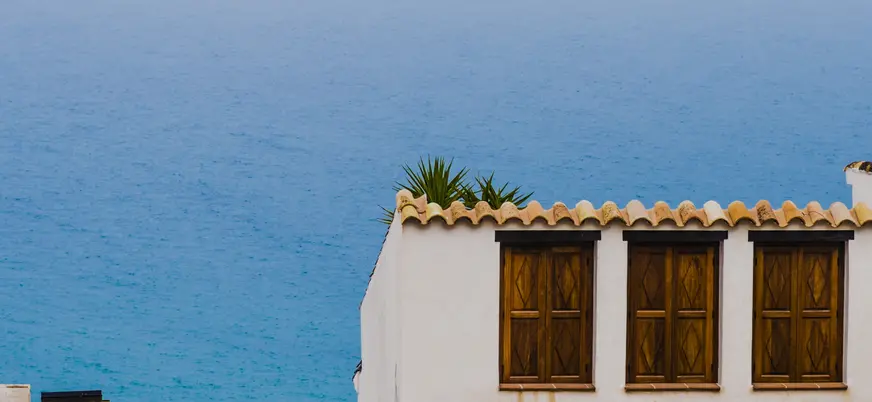 Fachada de casa mediterránea con tejado de tejas, ventanas de madera y el mar azul de fondo. Ideal para alquiler vacacional.