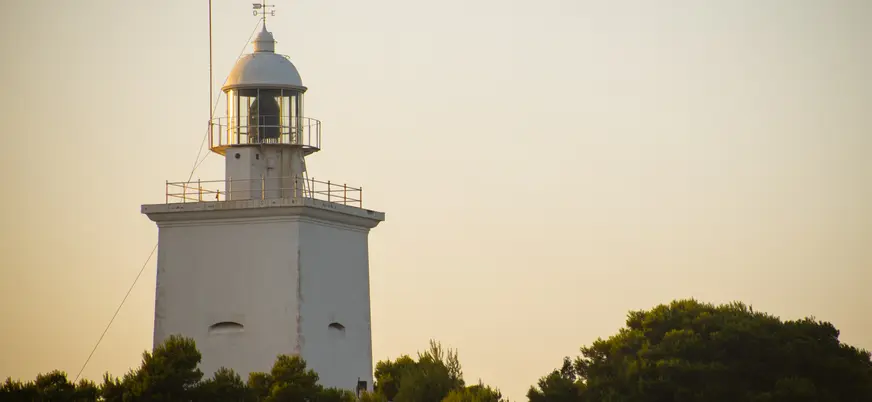 Faro blanco de Santa Pola, Alicante, destacando sobre un horizonte de color naranja pálido al amanecer, con árboles debajo.