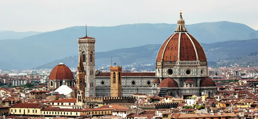 Vista panorámica de la Catedral de Santa María del Fiore en Florencia, Italia, destacando la cúpula de Brunelleschi, el campanario de Giotto y los tejados históricos de la ciudad con montañas al fondo.
