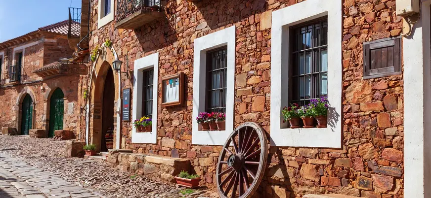 Calle de Castrillo de los Polvazares, León, con casa maragata de piedra rojiza, ventanas encaladas y rueda de carro.