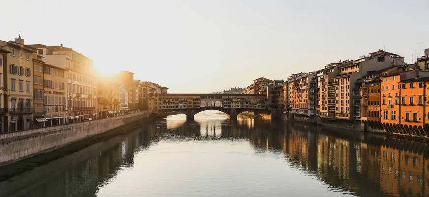 Vista del Ponte Vecchio en Florencia, Italia, al atardecer, con reflejos dorados en el río Arno y edificios históricos a ambos lados.
