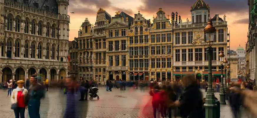 Personas paseando frente a edificios históricos con detalles dorados en la Grand Place de Bruselas al atardecer.