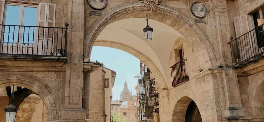 Arco de piedra en el centro histórico de Salamanca, con bustos y faroles colgantes, y la cúpula de la Catedral al fondo.