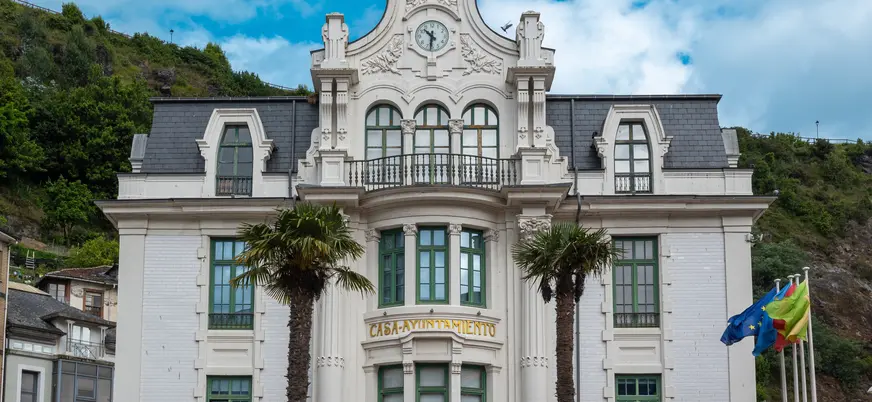 Fachada modernista de la Casa Ayuntamiento de Luarca, Asturias, con palmeras y reloj central.