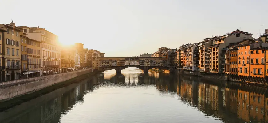 Vista del Ponte Vecchio en Florencia al atardecer, con el sol iluminando los edificios históricos a ambos lados del río Arno y reflejos en el agua.