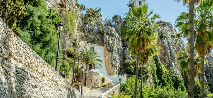 Escalinata y vegetación que llevan al castillo y campanario de Guadalest entre montañas.