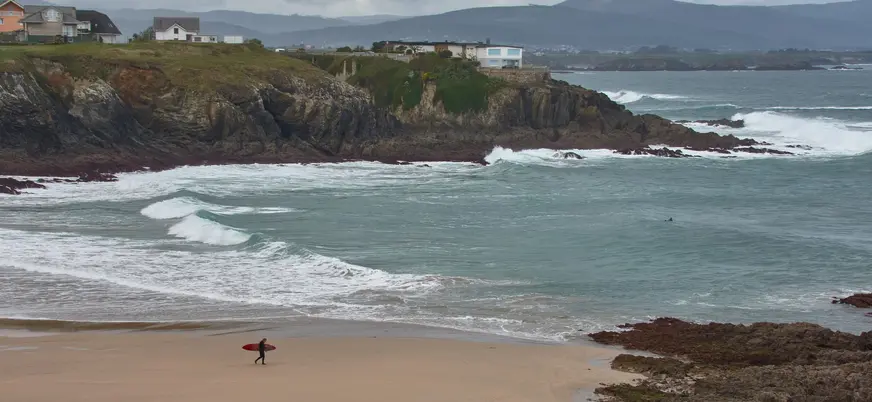 Surfista con tabla roja caminando por la playa de Tapia de Casariego, con el mar Cantábrico y acantilados al fondo.