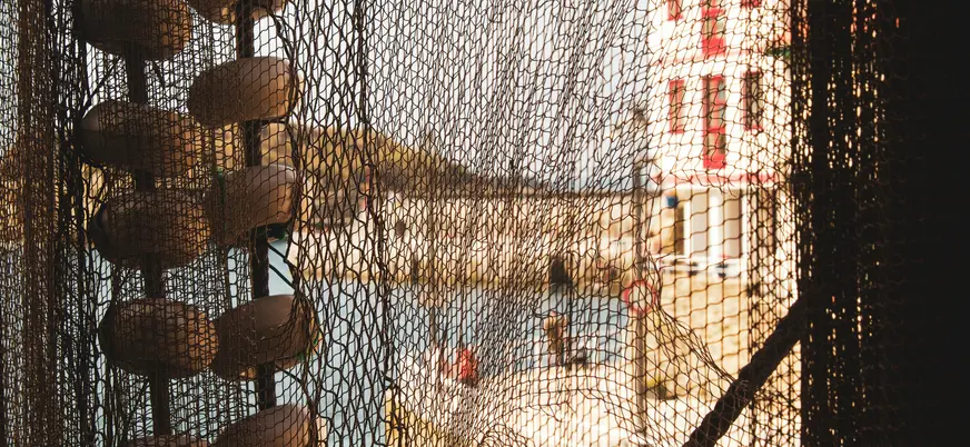 Detalle de redes y boyas de pesca en el puerto de Lastres, Asturias, con un edificio rojo al fondo.
