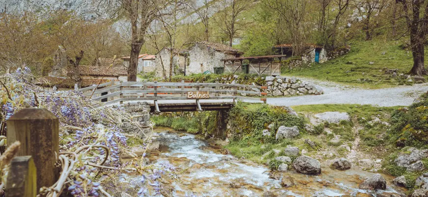 Puente de madera sobre el río Bulnes, en los Picos de Europa, rodeado de casas de piedra y un paisaje natural típico asturiano.