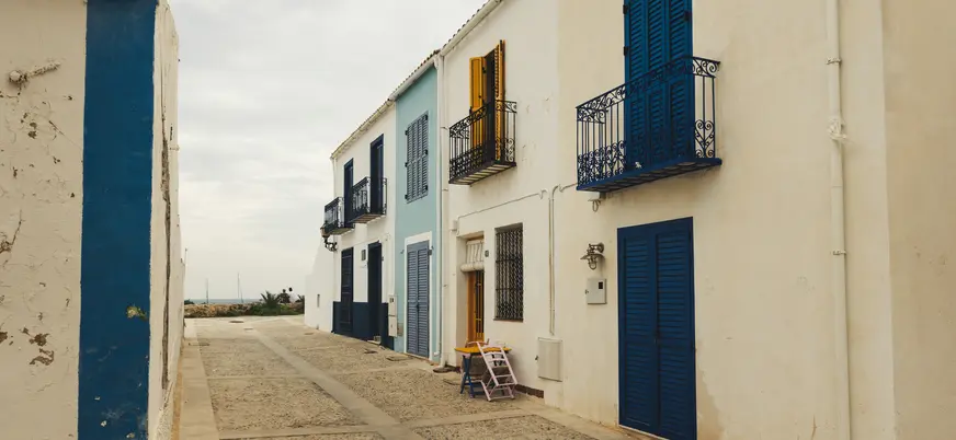 Calle empedrada de Tabarca con casas blancas y contraventanas azules y amarillas bajo un cielo nublado.