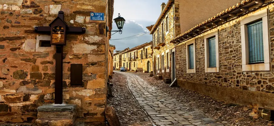 Calle empedrada tradicional en Castrillo de los Polvazares, León, con casas de piedra maragata y un crucero antiguo.