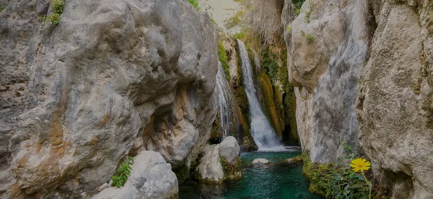 Cascada de las Fuentes del Algar en Alicante, cayendo entre grandes rocas hacia una poza de agua turquesa.