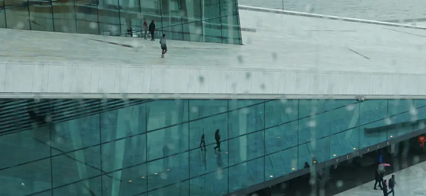 Reflejo en los cristales de la Ópera de Oslo, Noruega, con techo de mármol blanco y personas paseando bajo la lluvia. (