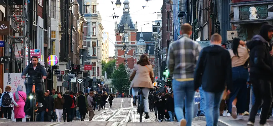Calle concurrida de Ámsterdam con personas caminando y ciclistas rodeada de edificios históricos y tiendas.