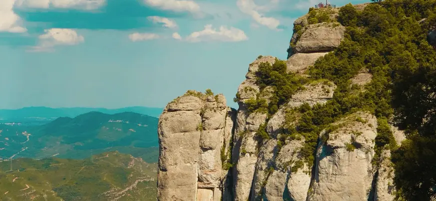 Peñasco de Montserrat con cruz en la cima y montañas verdes al fondo.