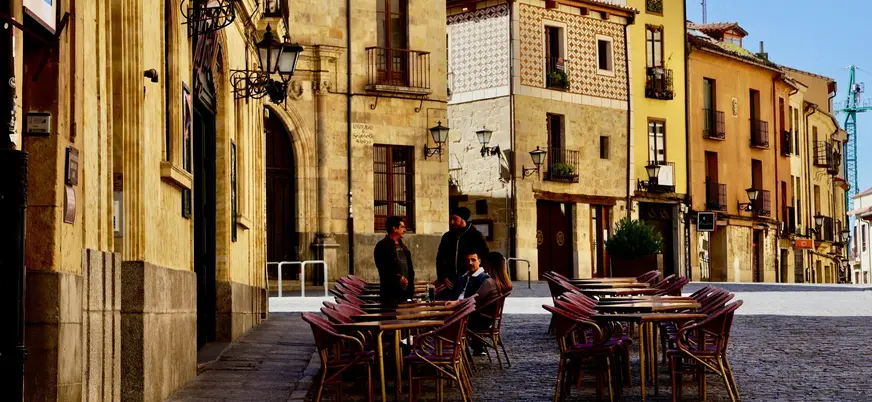 Terraza de café con mesas y sillas en una calle adoquinada del casco antiguo de Salamanca, edificios históricos y sol.
