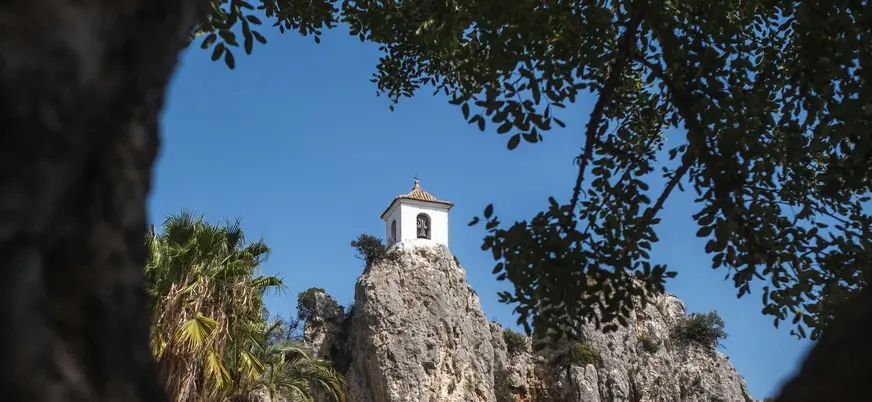 Campanario de Guadalest, un edificio blanco en la cima de una roca, visto a través de las ramas de los árboles.