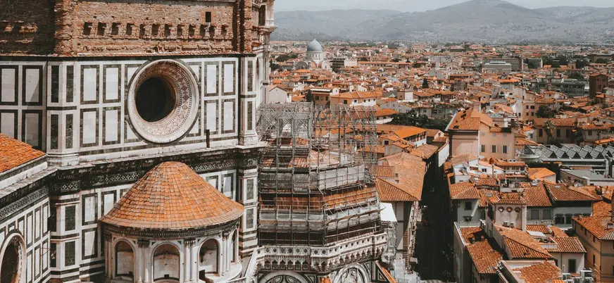 Vista detallada de la Catedral de Santa María del Fiore en Florencia, Italia, mostrando la fachada con mármol policromado y tejados de terracota, junto al paisaje urbano y montañas al fondo.