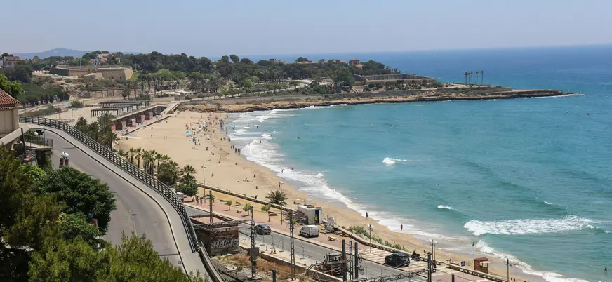 Vista aérea de la Playa del Miracle en Tarragona, con la línea costera, el mar azul y la carretera y tren en primer plano.