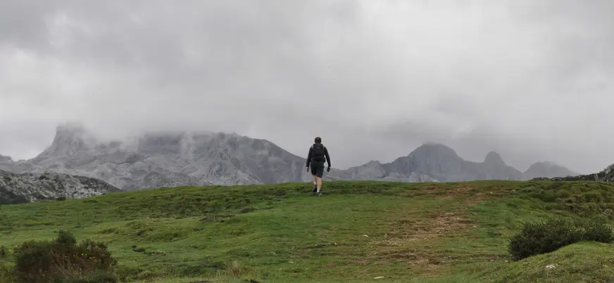 Persona caminando entre la niebla en los Lagos de Covadonga, Asturias, rodeada de montañas verdes y un cielo nublado.