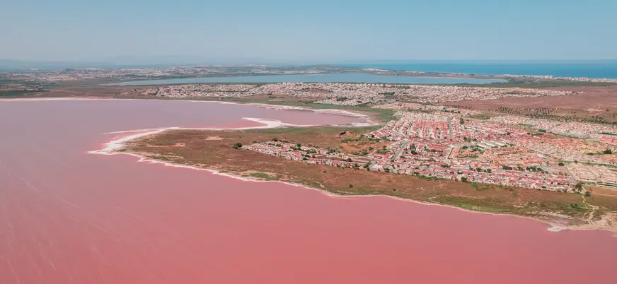 Vista aérea de la Laguna Rosa de Torrevieja con su intenso color rosa, zonas residenciales y la laguna azul de fondo.