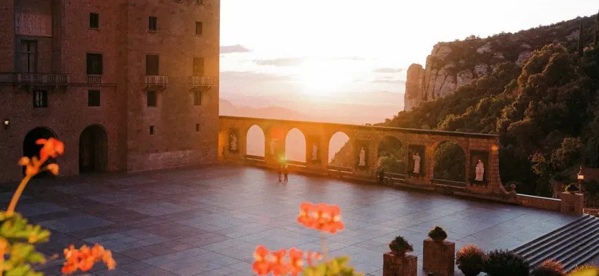 Patio del monasterio de Montserrat al atardecer, con arcos y montañas al fondo.