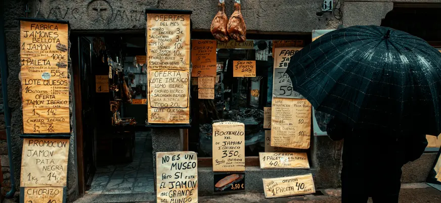 Tienda de jamón ibérico en España con ofertas escritas a mano y jamones colgando, persona con paraguas negro.