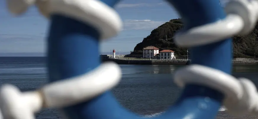 Vista del puerto de Ribadesella en Asturias, enmarcada por una barandilla azul con el faro y el mar Cantábrico al fondo.