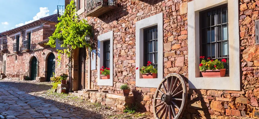 Calle empedrada de Castrillo de los Polvazares, León, con fachadas de piedra maragata y rueda de carro decorativa.