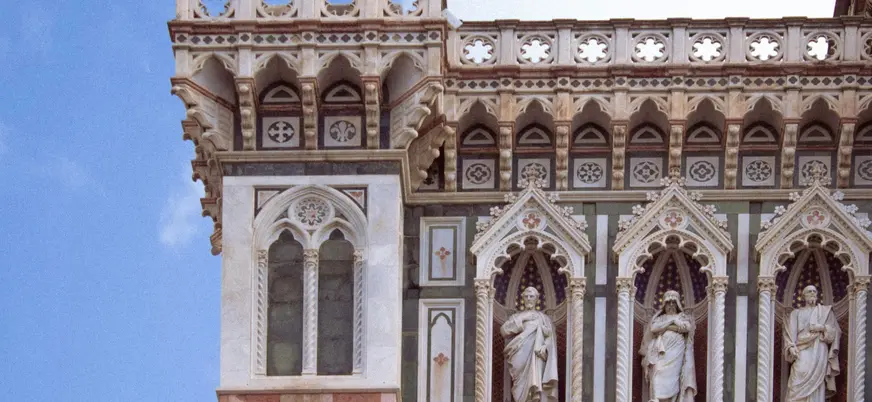 Detalle arquitectónico de la fachada del Duomo de Florencia, con ventanas góticas, esculturas religiosas en nichos y decoración ornamental en mármol blanco, verde y rosa.
