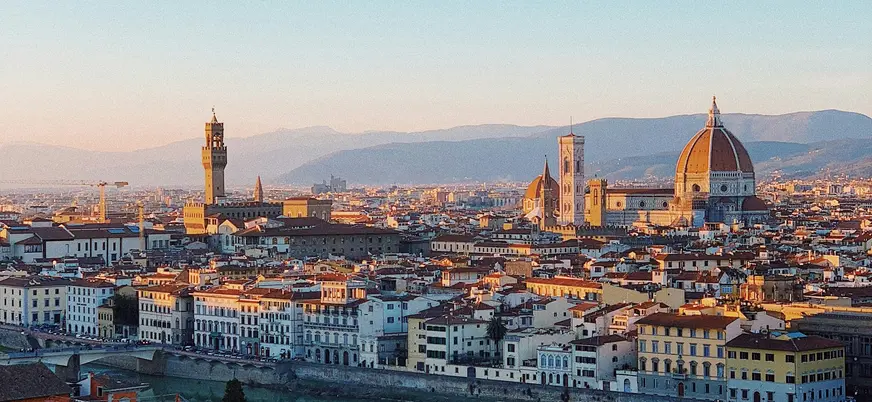 Vista panorámica de Florencia, Italia, con el río Arno en primer plano, la Catedral de Santa María del Fiore con su cúpula icónica, y la torre del Palazzo Vecchio al fondo, rodeados de edificios históricos y montañas.