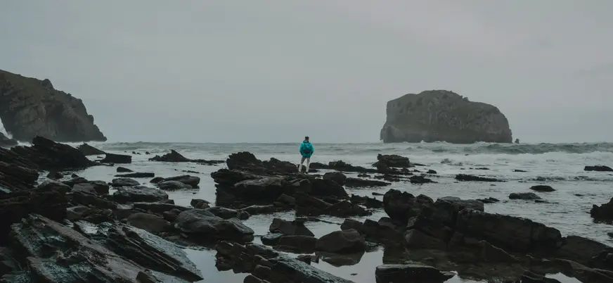 Persona sobre rocas frente al mar embravecido, con una isla rocosa al fondo bajo cielo gris.