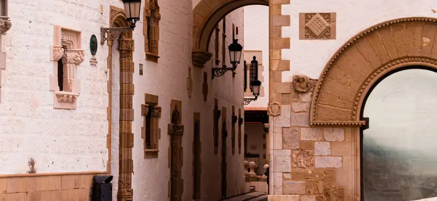 Calle estrecha de Sitges con muros blancos, arcos de piedra y faroles antiguos.