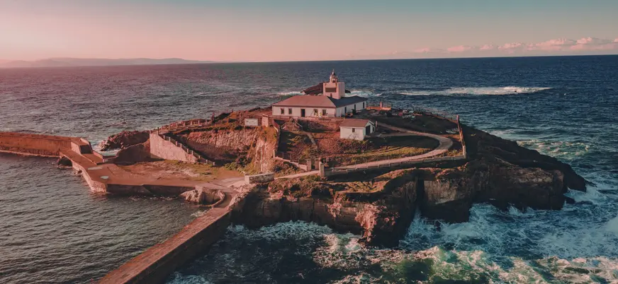 Vista aérea del faro de Tapia de Casariego al atardecer, rodeado por el mar Cantábrico y los acantilados de la costa asturiana.