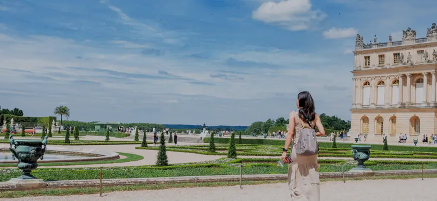 Una mujer con un vestido blanco camina hacia el Palacio de Versalles, mostrando su majestuosa fachada exterior.