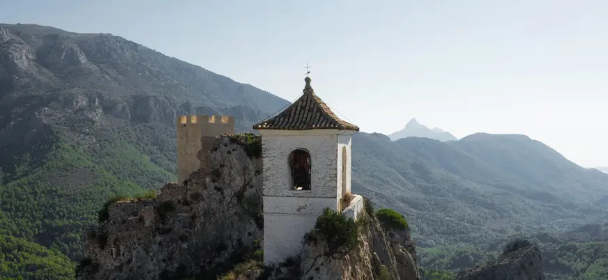 Vista aérea del campanario de Guadalest sobre un peñasco rocoso con montañas y vegetación de fondo.